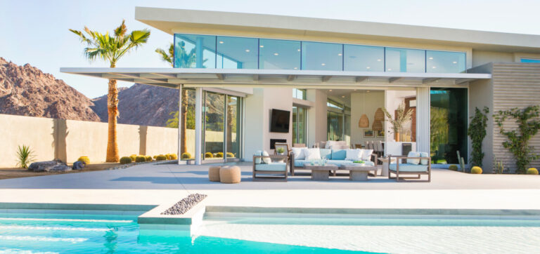 A modern desert home with pool and patio as seen from the backyard. The wall of sliding glass doors allows a glimpse inside.